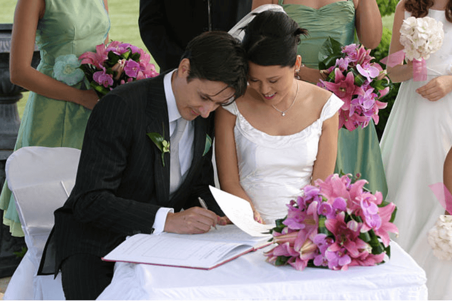 A newlywed couple sits at a table during their wedding ceremony, with the groom in a black pinstripe suit signing a document while the bride in a white wedding dress smiles beside him. Bridesmaids in pastel green dresses hold bouquets of pink and white flowers in the background. A bouquet of pink orchids rests on the table covered with a white cloth.