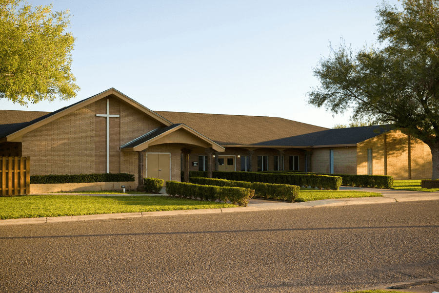 A modern church building with a brick exterior and a large white cross on the front stands under a clear sky. The well-manicured landscaping includes neatly trimmed hedges and green grass, with a paved road in the foreground. Trees cast long shadows, suggesting early morning or late afternoon sunlight.
