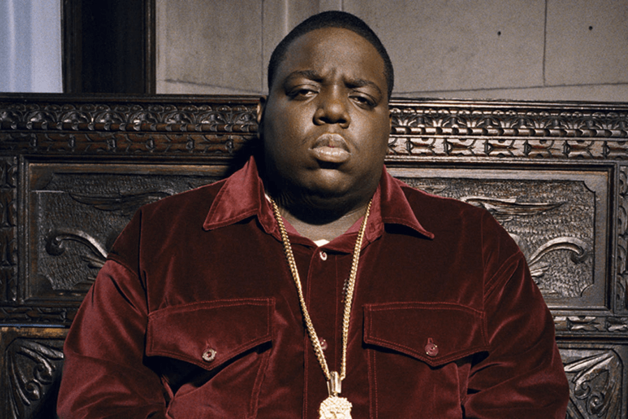 A man with a serious expression sits against an ornately carved wooden backdrop, wearing a deep red velvet shirt. He is accessorized with a thick gold chain featuring a large pendant, exuding a regal and powerful presence. The lighting casts soft shadows, emphasizing the richness of the fabric and the intricate details of the background.