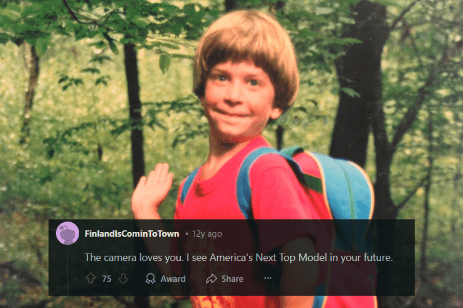 A young child with a bowl haircut poses dramatically in a forest setting, wearing a red t-shirt and a blue backpack. Their playful expression and exaggerated hand gesture give off an unintentional model-like vibe. The humorous caption below reads, "The camera loves you. I see America's Next Top Model in your future."