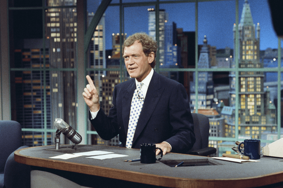 A man with curly brown hair, wearing a dark suit and a white shirt with a patterned tie, sits behind a wooden desk on a late-night talk show set. He gestures with one finger raised while speaking, with a classic microphone, papers, and coffee mugs on the desk. The background features a large window displaying a nighttime city skyline with tall buildings.