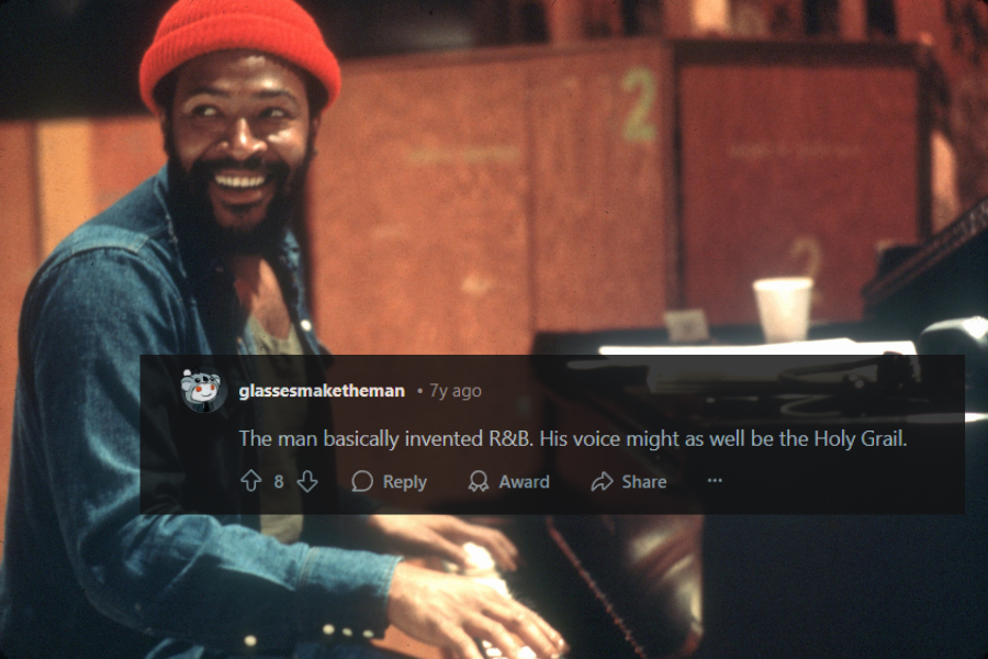 A male musician sits at a grand piano, smiling warmly while playing. He wears a red beanie, a denim jacket over a dark tank top, and has a neatly groomed beard. The setting appears to be a recording studio, with numbered soundproof panels and scattered papers on the piano.