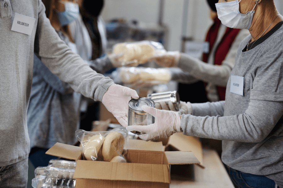 A group of volunteers wearing masks and gloves sorts and distributes food at a community donation center. One volunteer hands a canned food item to another, while boxes filled with bread and other supplies are stacked on a table. The volunteers wear name tags, and the scene conveys teamwork and generosity.