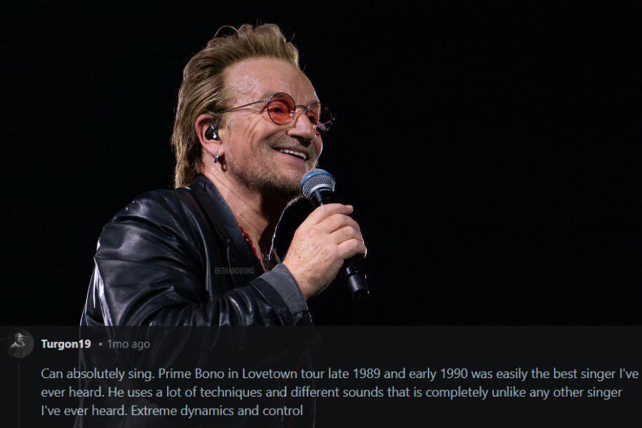 A male rock singer performs on stage, smiling while holding a microphone. He wears a black leather jacket, a red beaded necklace, and signature tinted glasses. The dark background highlights his expressive face and charismatic stage presence.