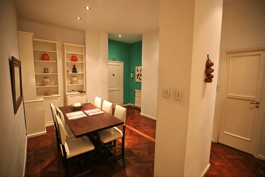 A warmly lit dining room with a dark wooden table and six cream-colored chairs, set against white walls and a parquet floor. Built-in shelves display decorative items, while a teal accent wall in the background contrasts with the neutral tones. A doorway and a hallway lead to other parts of the home.