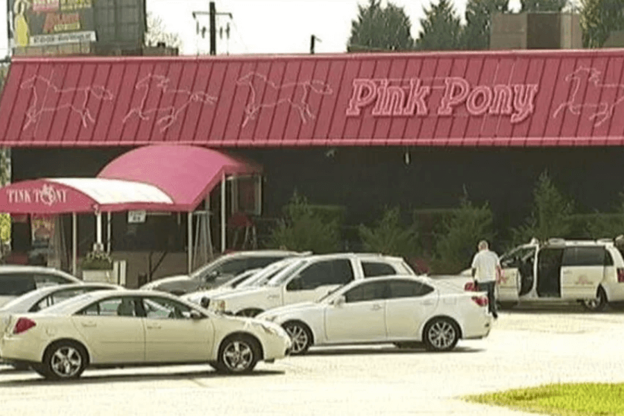 A daytime view of the exterior of the Pink Pony, a strip club with a red roof featuring neon horse silhouettes and a pink awning over the entrance. Several cars, including a white van with the club’s logo, are parked in the lot, and a man in a white shirt is standing near an open vehicle door. The surrounding area includes trees and a billboard in the background.