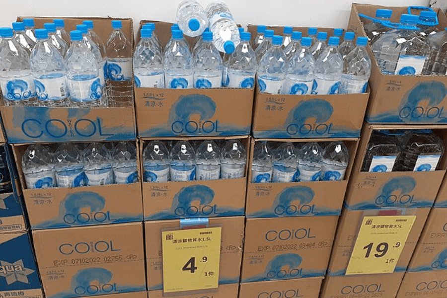 Stacks of cardboard boxes filled with "COOL" brand bottled water in a supermarket. The clear plastic bottles have blue caps and labels featuring a water-themed design. Yellow price tags indicate different prices for single bottles and bulk packs.