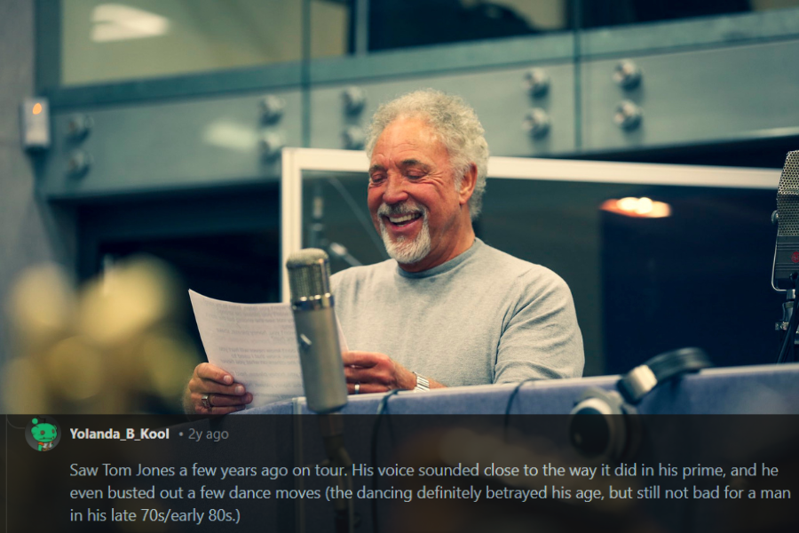 A male singer with curly gray hair and a beard laughs while holding a sheet of lyrics in a professional recording studio. He wears a casual gray sweater, standing behind a studio microphone with headphones resting on the booth. The warm lighting and blurred foreground elements create an intimate and joyful atmosphere.