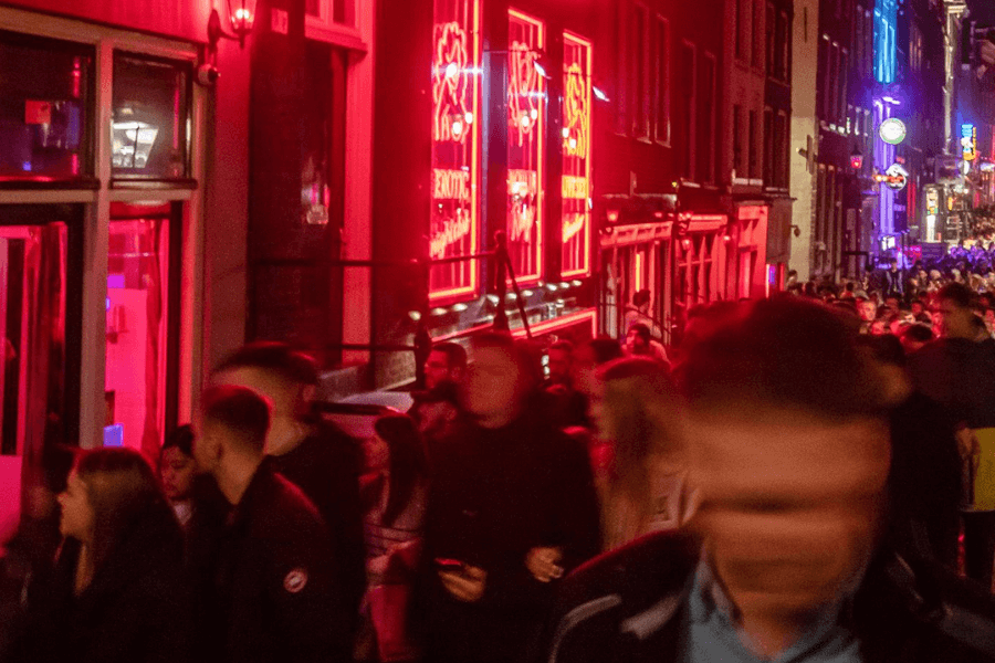 A crowded street at night illuminated by bright red neon signs advertising exotic entertainment and adult venues. The blurred movement of pedestrians creates a sense of bustling activity, with people walking past storefronts featuring glowing windows. The background extends into a lively nightlife district with additional colorful lights and signs.