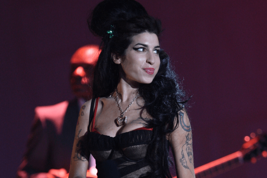 A female singer with a signature beehive hairstyle and winged eyeliner performs on stage, wearing a black and red corset-style dress. Her arms are adorned with tattoos, and she has a playful expression with her tongue slightly sticking out. A musician in a suit and a guitar are visible in the dimly lit background.