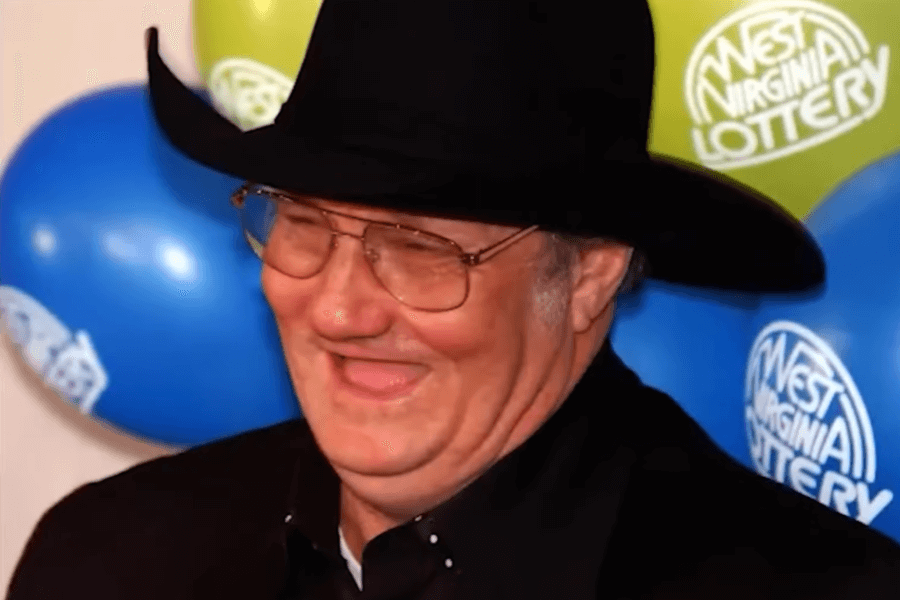 A man wearing a black cowboy hat and glasses is seen laughing with a joyful expression. He is dressed in a dark suit, and the background features blue and green balloons with the "West Virginia Lottery" logo.