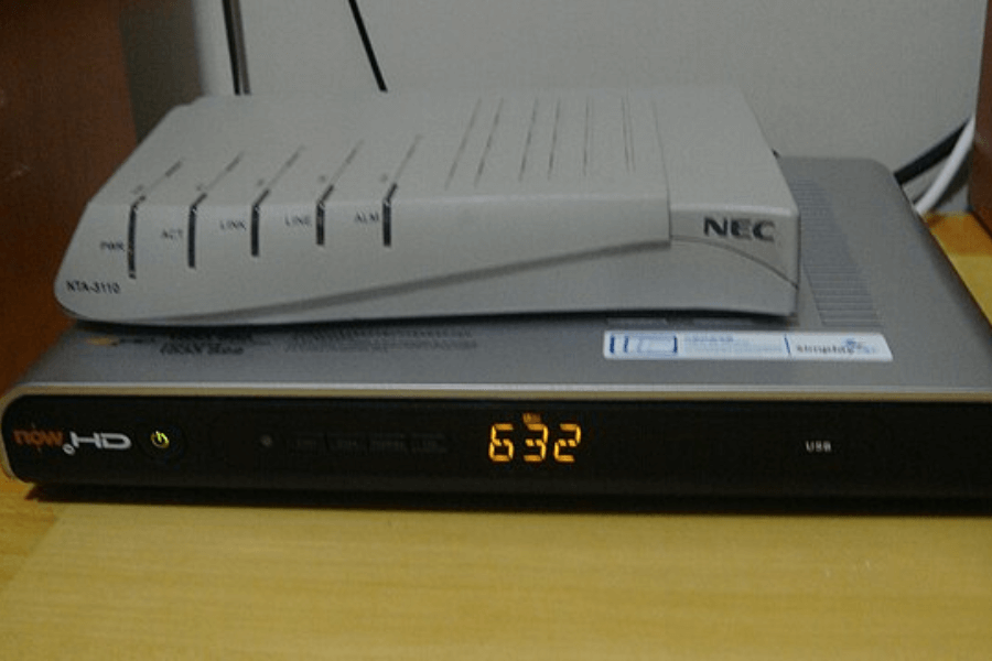 A grey NEC NTA-3110 network device with indicator lights is stacked on top of a Now HD set-top box with an illuminated digital clock displaying "6:32." The devices are placed on a wooden surface, with cables running in the background.