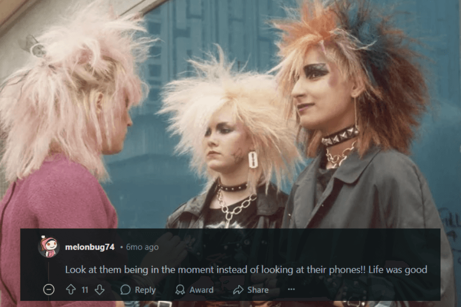 A group of three punk-styled individuals with extreme, teased, and colorful hairstyles stand together in an urban setting, engaged in conversation. Their outfits include leather jackets, studded accessories, bold makeup, and spiked chokers, embodying the rebellious punk aesthetic of the 1980s. A caption below humorously comments on how they are “being in the moment” instead of looking at their phones, reminiscing about a simpler time.