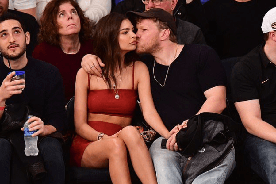 A woman in a red two-piece outfit sits courtside at a sports event while a man in a black shirt and cap leans in to kiss her cheek. She looks away with a neutral expression, her arms resting on her lap. Surrounding them, other spectators are engaged in various activities, including a man holding a water bottle and a woman looking up with a curious expression.