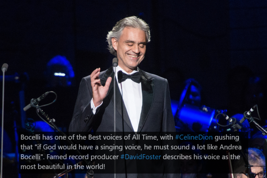 A male classical singer in a black tuxedo and bow tie stands on stage, smiling with his eyes closed while performing. He gestures gracefully with one hand near the microphone, exuding elegance and emotion. The dimly lit background features an orchestra with musicians illuminated by blue stage lighting.