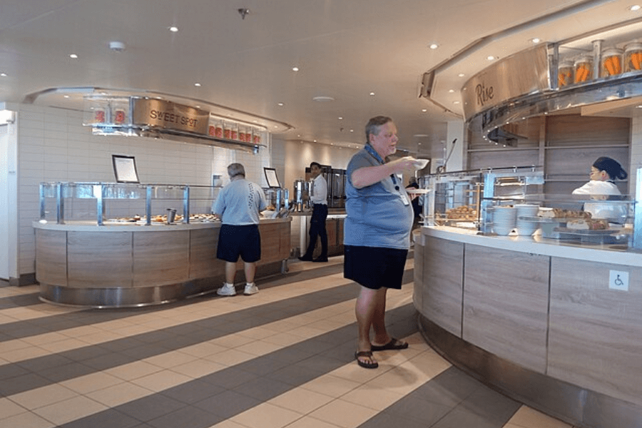 A spacious cruise ship buffet with modern wooden and glass serving stations labeled "Sweet Spot" and "Rise." Guests in casual clothing are selecting food, while a staff member behind the counter serves baked goods. The interior is well-lit with a clean, contemporary design featuring tiled floors and stainless steel accents.