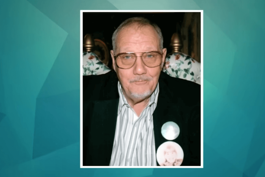 A framed photograph of an elderly man with glasses and a trimmed beard, wearing a black blazer over a striped button-down shirt. He has a neutral expression and is sitting in a chair with a floral-patterned cushion in the background. Two round pins are attached to his blazer, one featuring a faded image of a person. The image is displayed on a teal-colored digital background.
