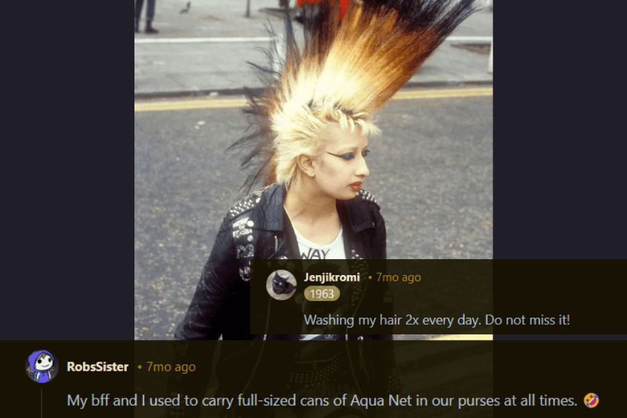 A punk woman with a towering mohawk, featuring a striking mix of bleached blonde and black tips, stands on a city street. She wears a studded leather jacket over a graphic t-shirt, fully embracing the rebellious punk aesthetic. The comments below joke about the maintenance of such a hairstyle, mentioning washing hair twice a day and carrying full-sized cans of Aqua Net hairspray everywhere.