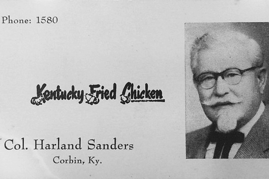 A vintage black and white business card for Kentucky Fried Chicken, featuring a small photograph of Colonel Harland Sanders wearing his signature glasses, mustache, and bow tie. The card includes the text "Phone: 1580," "Kentucky Fried Chicken" with decorative chicken illustrations, and "Col. Harland Sanders, Corbin, Ky."