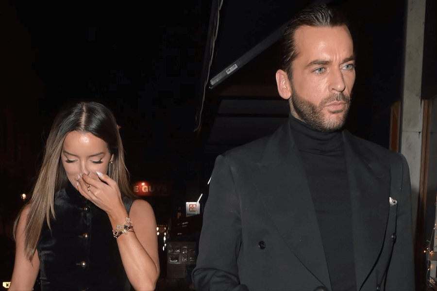 A well-dressed man in an all-black outfit, including a tailored blazer and turtleneck, walks with a serious expression on a dimly lit street at night. Beside him, a woman in a sleeveless black velvet dress covers her mouth with her hand, possibly laughing or hiding her face. The background features streetlights and a faintly visible sign, suggesting a city nightlife setting.
