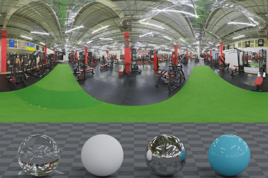 A 360-degree panoramic image of a spacious gym with black flooring, red support columns, and a green turf strip running through the center. Various strength training and cardio equipment are neatly arranged, and the ceiling is covered with reflective insulation. At the bottom of the image, four material test spheres—glass, matte white, chrome, and glossy blue—are placed on a checkered surface.