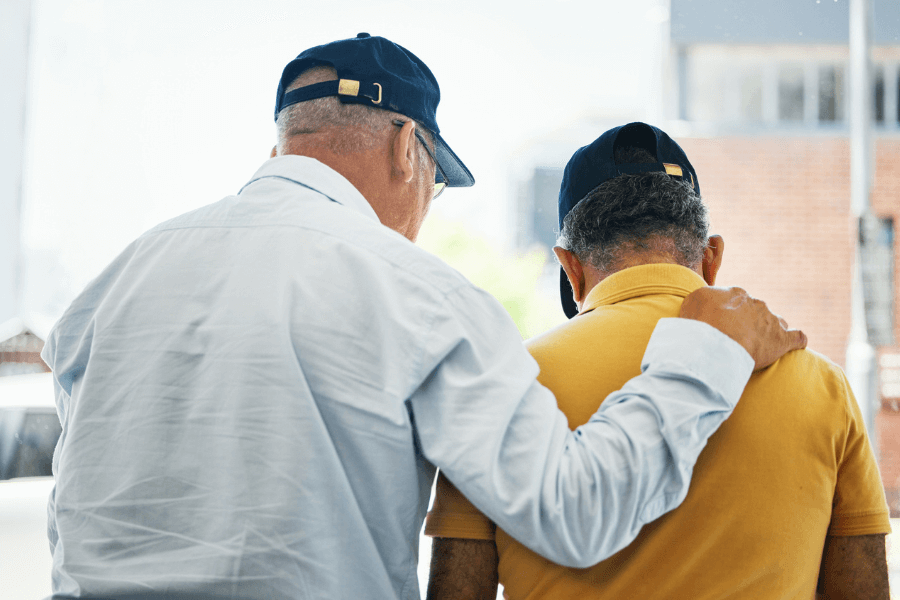 Two elderly men wearing baseball caps stand side by side, with one placing a comforting hand on the other's shoulder. One man wears a white button-up shirt, while the other is dressed in a yellow polo shirt. The background is blurred, showing an outdoor urban setting with buildings and street elements.