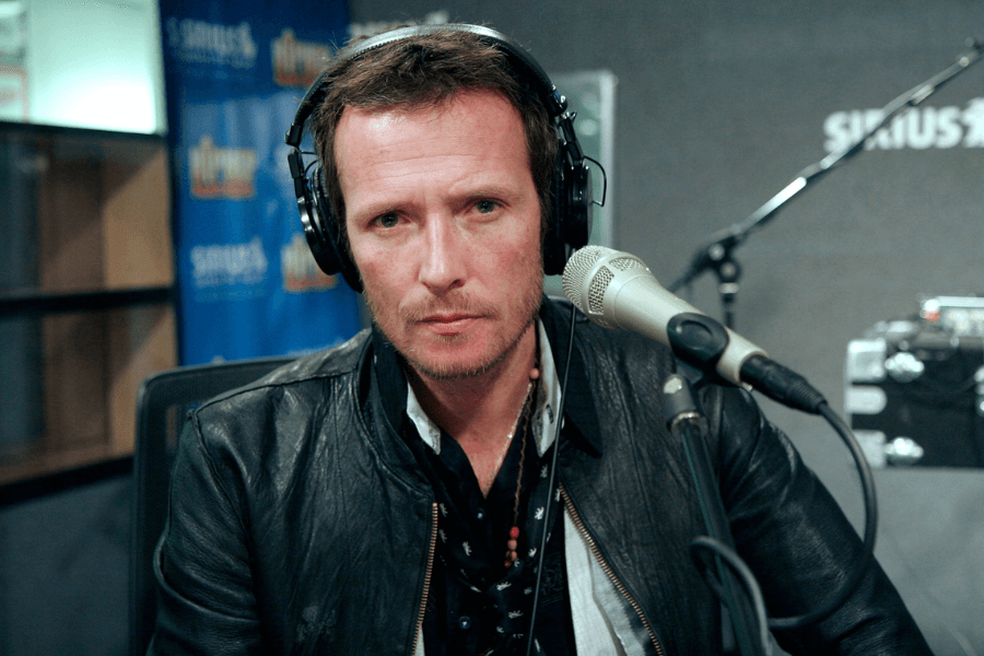 A male musician with short, slightly unkempt hair and a serious expression sits in a radio studio wearing large over-ear headphones. He is dressed in a black leather jacket, a patterned scarf, and a beaded necklace, leaning slightly toward a professional microphone. The "SiriusXM" logo and recording equipment are visible in the background, emphasizing the broadcast setting.