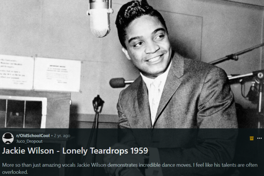 A black and white photograph of a male singer in a recording studio, smiling as he leans on a microphone. He is dressed in a sharp suit with a neatly styled pompadour, exuding charm and confidence. The vintage studio equipment and warm expression capture a timeless moment in music history.