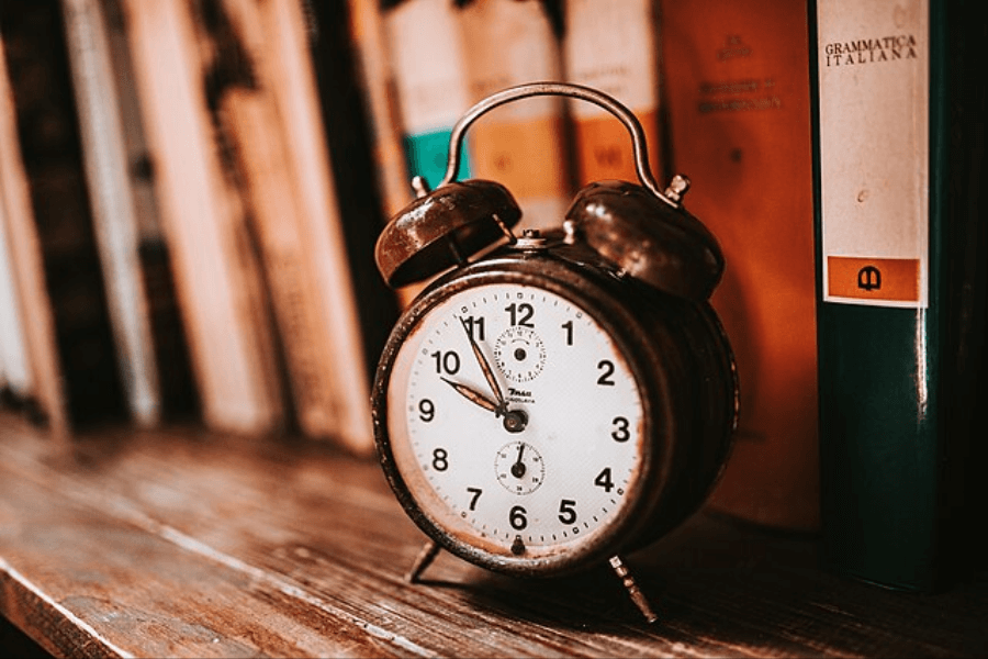 A vintage, rusted alarm clock with a white face and black hands sits on a wooden shelf, showing the time as approximately 10:10. Behind it, a row of old books, including one titled "Grammatica Italiana," is slightly out of focus. The scene has a warm, nostalgic ambiance with soft lighting.