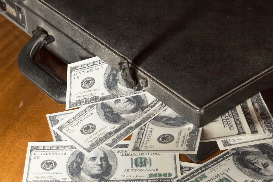 A black briefcase sits partially open on a wooden surface, with numerous $100 bills spilling out. The crisp banknotes, featuring Benjamin Franklin’s portrait, suggest a large sum of cash. The lighting casts shadows, emphasizing the texture of the case and money.