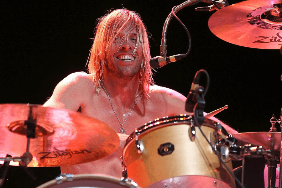 A shirtless drummer with long, blonde, sweat-soaked hair energetically plays his drum kit during a live performance. He is smiling broadly, with a microphone positioned close to his mouth, possibly singing while drumming. The stage lighting reflects off the cymbals, highlighting the excitement and intensity of the moment.