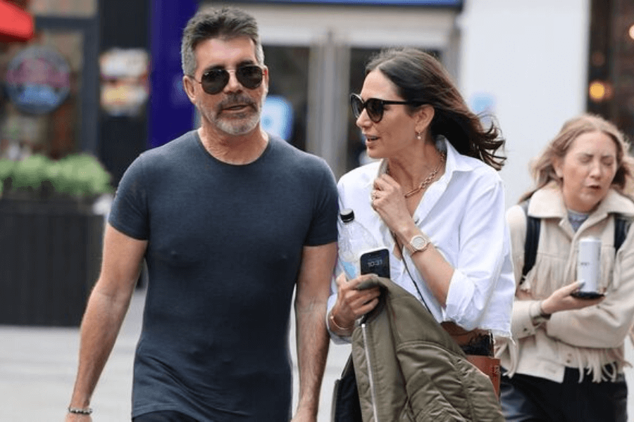A man with short, dark hair, a beard, and sunglasses walks through a city street wearing a tight-fitting dark gray T-shirt. Beside him, a woman in a white button-up shirt, sunglasses, and gold jewelry walks while speaking, holding a green jacket and a water bottle. The background features other pedestrians, including a woman in a beige coat drinking from a can.