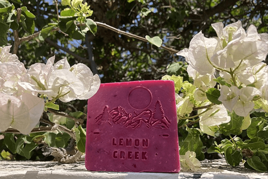 A vibrant pink handcrafted soap bar with "LEMON CREEK" embossed along with a mountain and tree design sits on a sunlit surface. White bougainvillea flowers frame the soap, with green foliage in the background, creating a natural, outdoor setting.