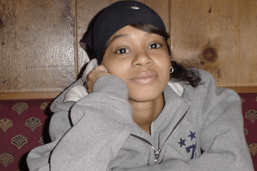 A young woman with warm eyes and a relaxed expression rests her head on her hand while sitting in a cozy booth. She wears a black bandana, silver hoop earrings, and a gray zip-up hoodie with star details on the chest. The wooden-paneled background and patterned seating create a casual, intimate atmosphere.