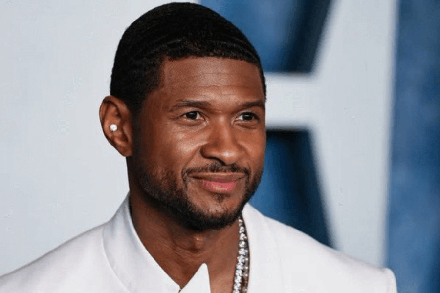 A man with neatly trimmed facial hair and short, wavy hair smiles subtly while dressed in a white suit with a diamond chain necklace. He wears a stud earring and has a confident yet relaxed expression. The blurred blue and white background suggests a formal or red carpet event.
