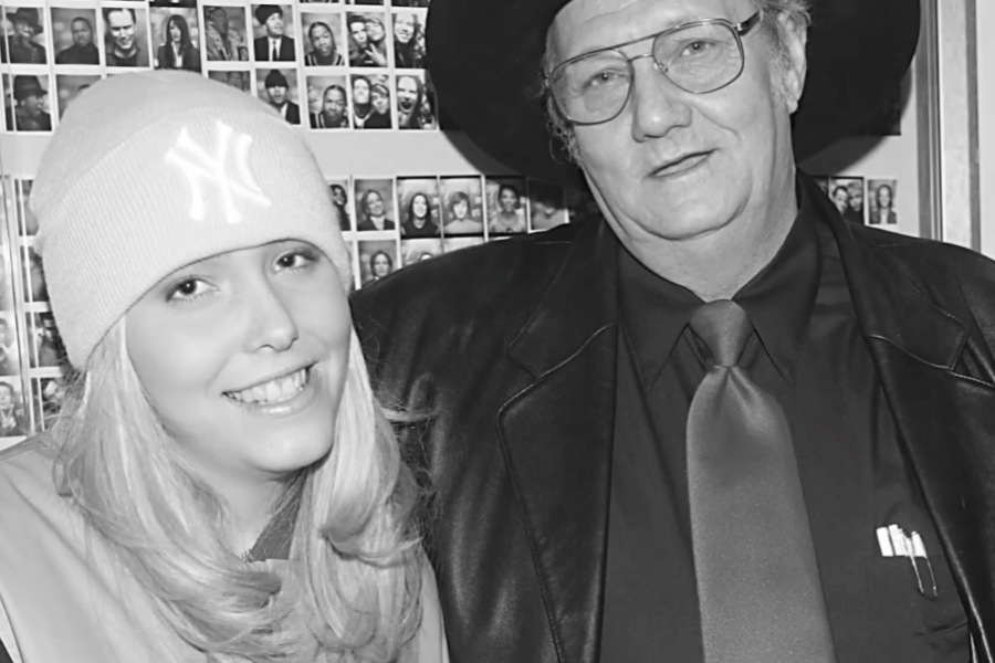 A black and white photograph of a young blonde woman wearing a New York Yankees beanie and a smiling expression, standing next to an older man in a black cowboy hat, glasses, and a leather jacket. The man wears a dark shirt and tie, with pens visible in his jacket pocket. Behind them, a wall covered in small photographs of people is visible.