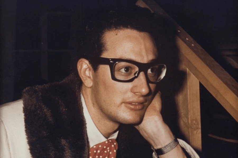 A young man with thick black-framed glasses and neatly combed hair leans on his hand with a thoughtful expression. He wears a white suit jacket with a fur collar, a red polka-dotted bow tie, and a silver wristwatch. The warm lighting and vintage setting create a nostalgic, classic feel.