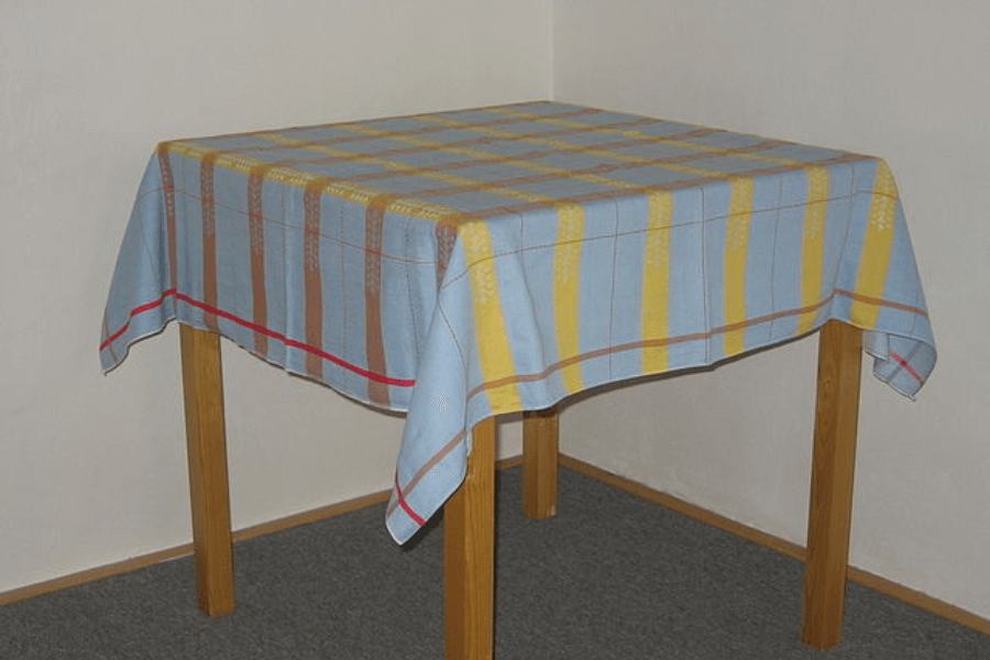 A simple wooden table covered with a checkered tablecloth featuring blue, yellow, and brown stripes with a red border. The table is set against a plain white wall, with a grey carpeted floor beneath it. The fabric drapes evenly over the edges of the table.