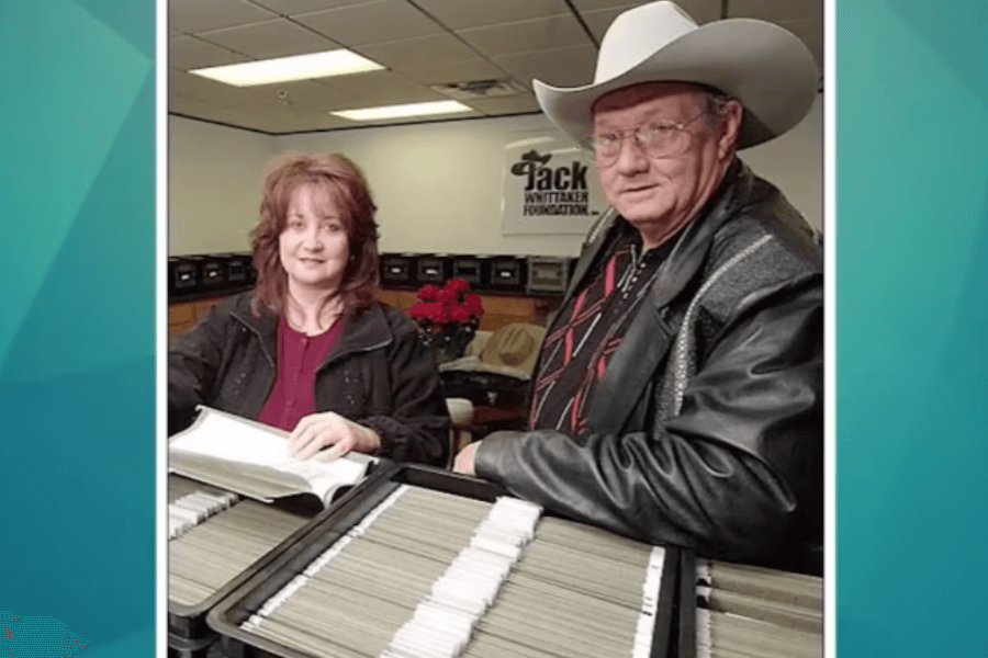 A man in a white cowboy hat, glasses, and a black leather jacket stands next to a woman with shoulder-length red hair, both looking at documents inside an office. The room has filing boxes neatly arranged along the walls, and a sign in the background reads "Jack Whittaker Foundation." The man appears to be engaged in paperwork, while the woman smiles slightly at the camera.
