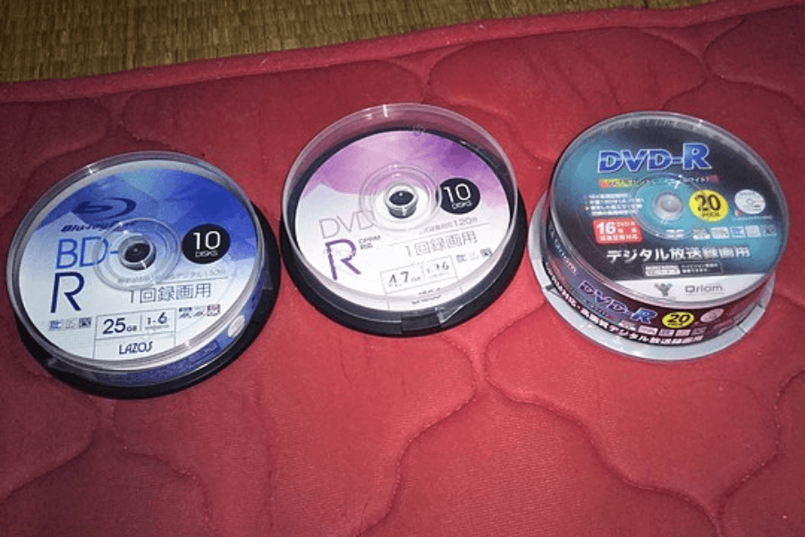 Three spindles of blank recordable discs—one Blu-ray BD-R, one DVD-R with a purple label, and another DVD-R with a blue label—placed on a red quilted surface. Each spindle contains multiple discs, labeled with storage capacities and recording speeds, with text in Japanese.