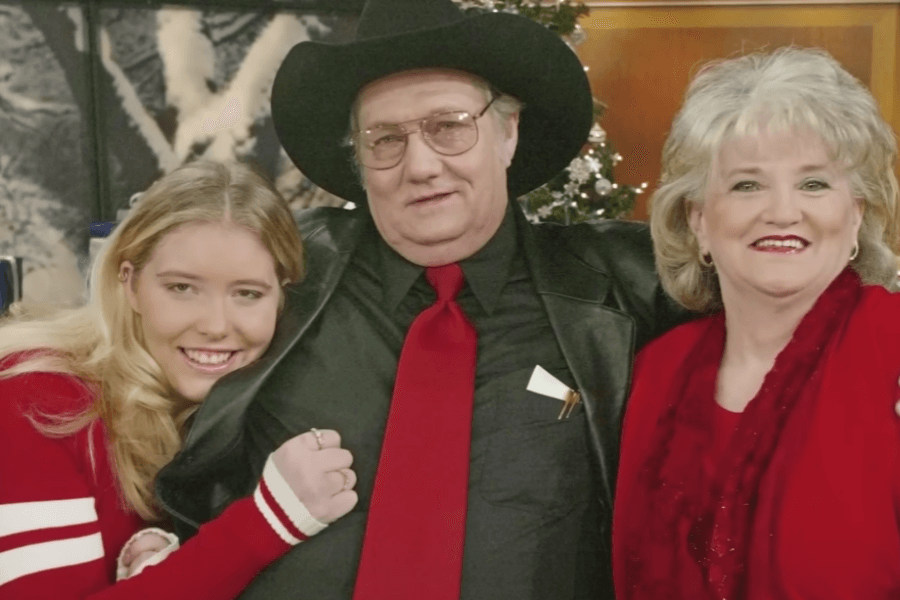 A man in a black cowboy hat, glasses, and a black leather jacket with a red tie stands between two smiling women dressed in red. The young blonde woman on the left leans in, holding onto his arm, while the older woman on the right beams at the camera. A decorated Christmas tree and festive winter-themed backdrop are visible in the background.