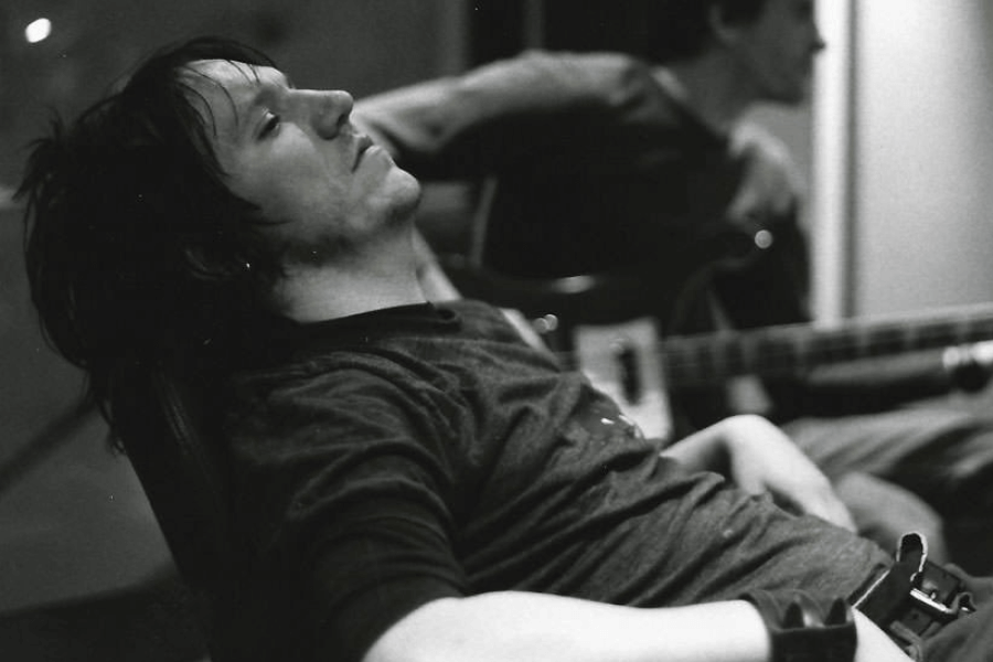 A black-and-white photograph of a young man with messy dark hair leaning back in a chair, eyes closed, with a contemplative expression. He wears a casual, slightly wrinkled t-shirt, and a leather wristband is visible on his arm. In the blurred background, another musician is playing a bass guitar, adding to the moody, intimate atmosphere of the scene.