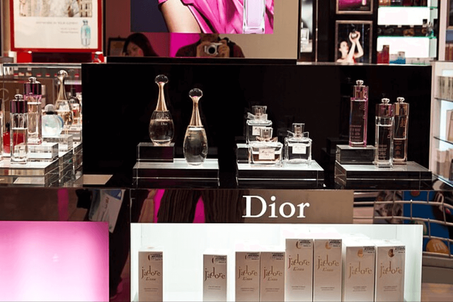 A luxury Dior perfume display in a retail store showcasing an array of fragrances, including J’adore and other signature scents in elegant glass bottles. The display features a black and mirrored background, with a lower shelf holding neatly arranged J’adore perfume boxes. A reflection of a person taking a photo is visible in the mirror.