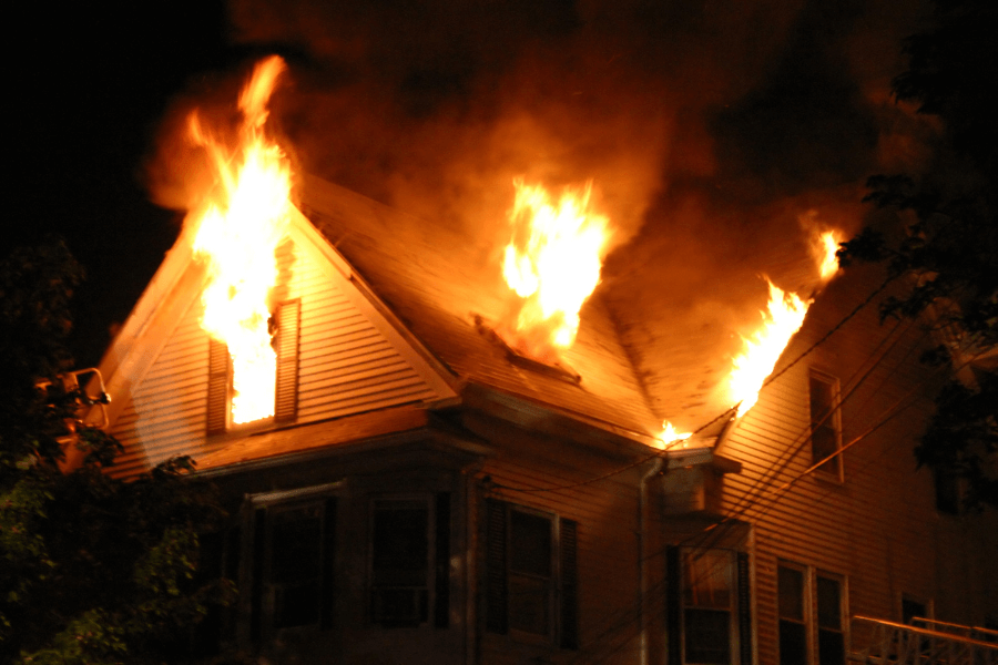 A large two-story house is engulfed in flames at night, with fire bursting out from the roof and upper windows. Thick black smoke rises into the dark sky, and the intense glow of the fire illuminates the surrounding area. Power lines and trees are visible near the burning structure.