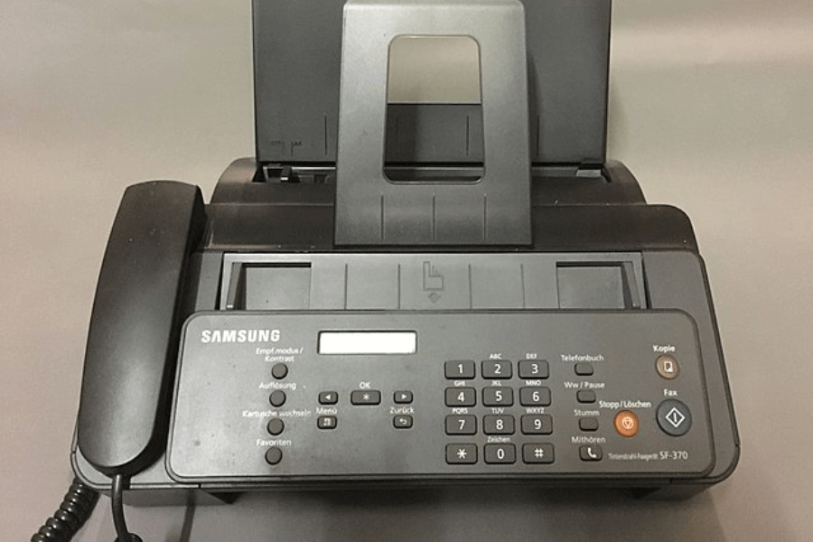 A Samsung SF-370 fax machine with a built-in telephone handset, numeric keypad, and function buttons. The machine is black with a paper feeder on top and a small digital display for settings and controls. It is placed on a neutral-colored surface.