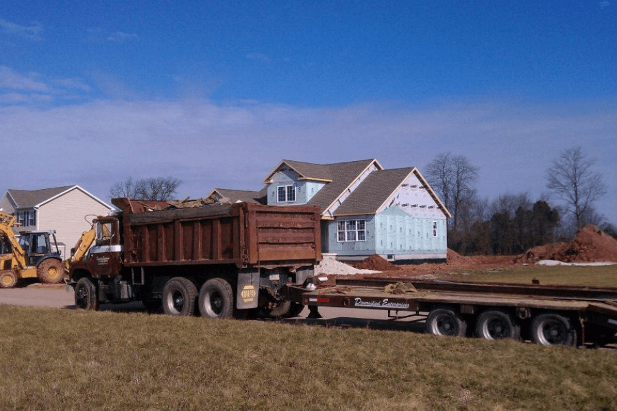 A construction site with a partially built house covered in blue insulation panels, surrounded by dirt piles and construction materials. In the foreground, a rusted dump truck with a flatbed trailer labeled "Diversified Enterprises" is parked on a grassy field. Several yellow backhoes and other construction equipment are visible in the background near completed homes under a clear blue sky.
