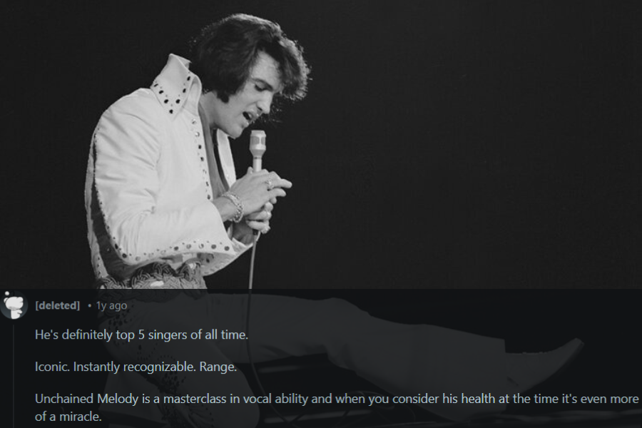 A black and white photograph of a male singer mid-performance, dressed in an embellished white jumpsuit with a high collar and a wide belt. He holds a microphone close to his mouth, singing with intensity while extending one leg outward in a dramatic pose. The dark background emphasizes his stage presence and iconic style.