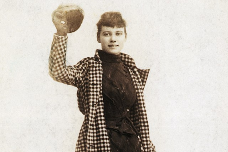 A vintage sepia-toned photograph of a woman wearing a checkered coat over a dark dress, holding up a round hat with a confident smile. Her short, wavy hair and relaxed pose give her an adventurous and independent demeanor. The plain background emphasizes her as the focal point of the image.