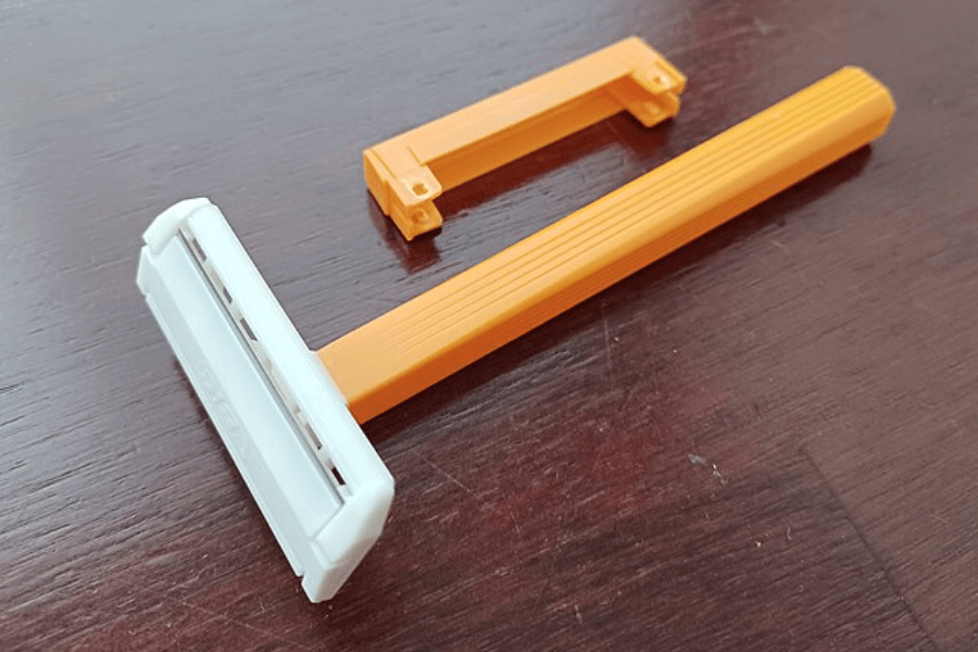 A simple disposable razor with a bright orange handle and a white blade cartridge resting on a dark wooden surface. Next to it is a detached orange plastic cover designed to protect the razor blades.