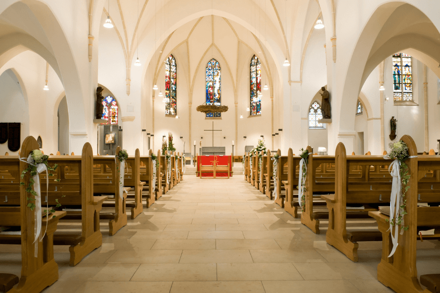 A beautifully decorated church interior with wooden pews adorned with white ribbons and fresh greenery, set up for a wedding ceremony. The high, arched ceilings and elegant stained-glass windows allow natural light to illuminate the space. The altar at the front is covered in red cloth, with a simple cross as the focal point.